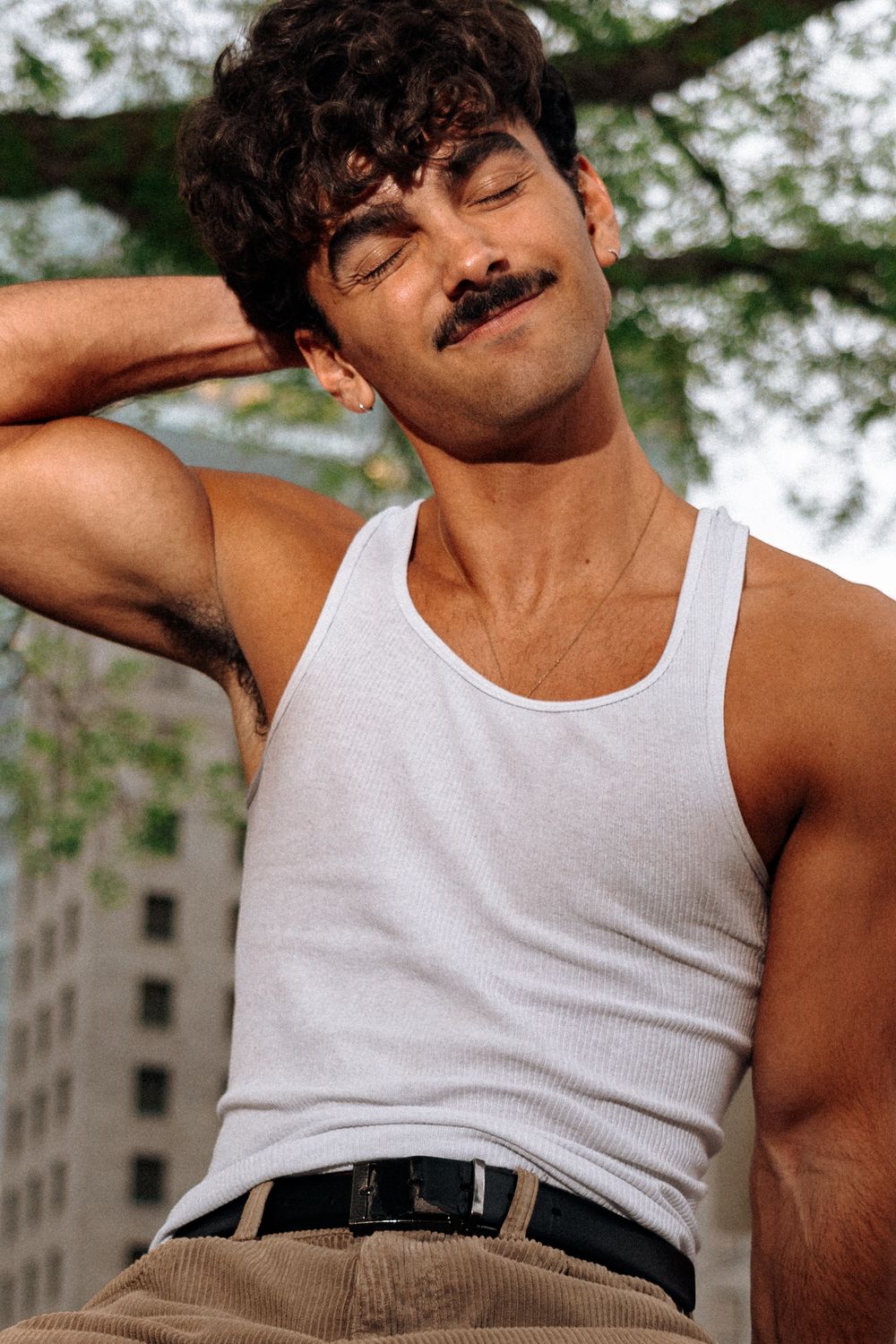 Young man with mustache in white tank top posing outdoors with eyes closed, hand behind neck.