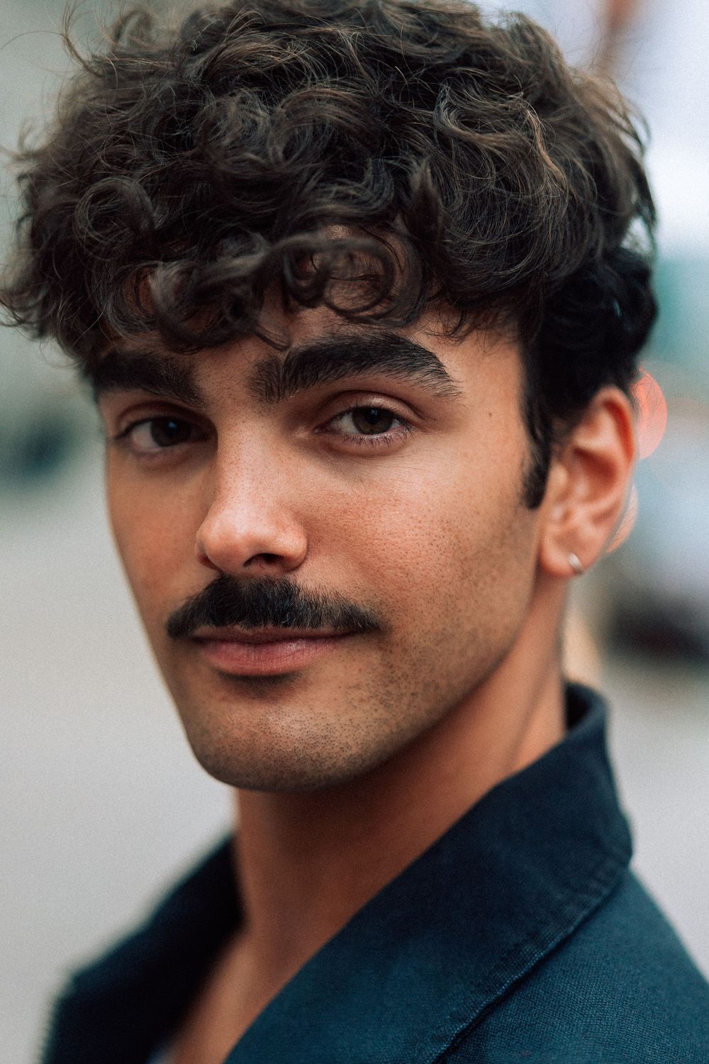 Young man with curly dark hair and mustache wearing a teal jacket, posing outdoors in a close-up portrait.