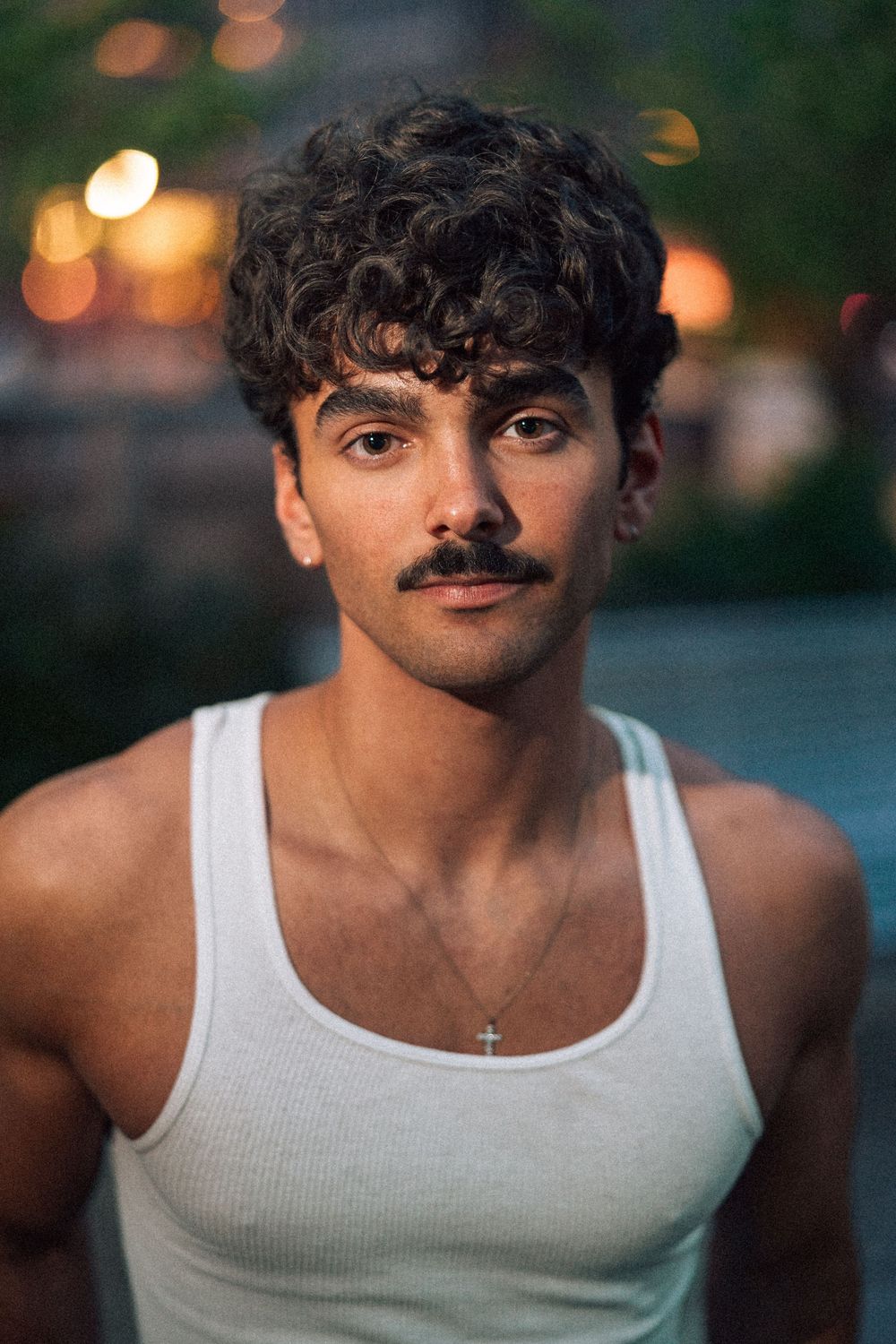 Young man with curly hair and mustache wearing a white tank top and cross necklace outdoors at dusk.