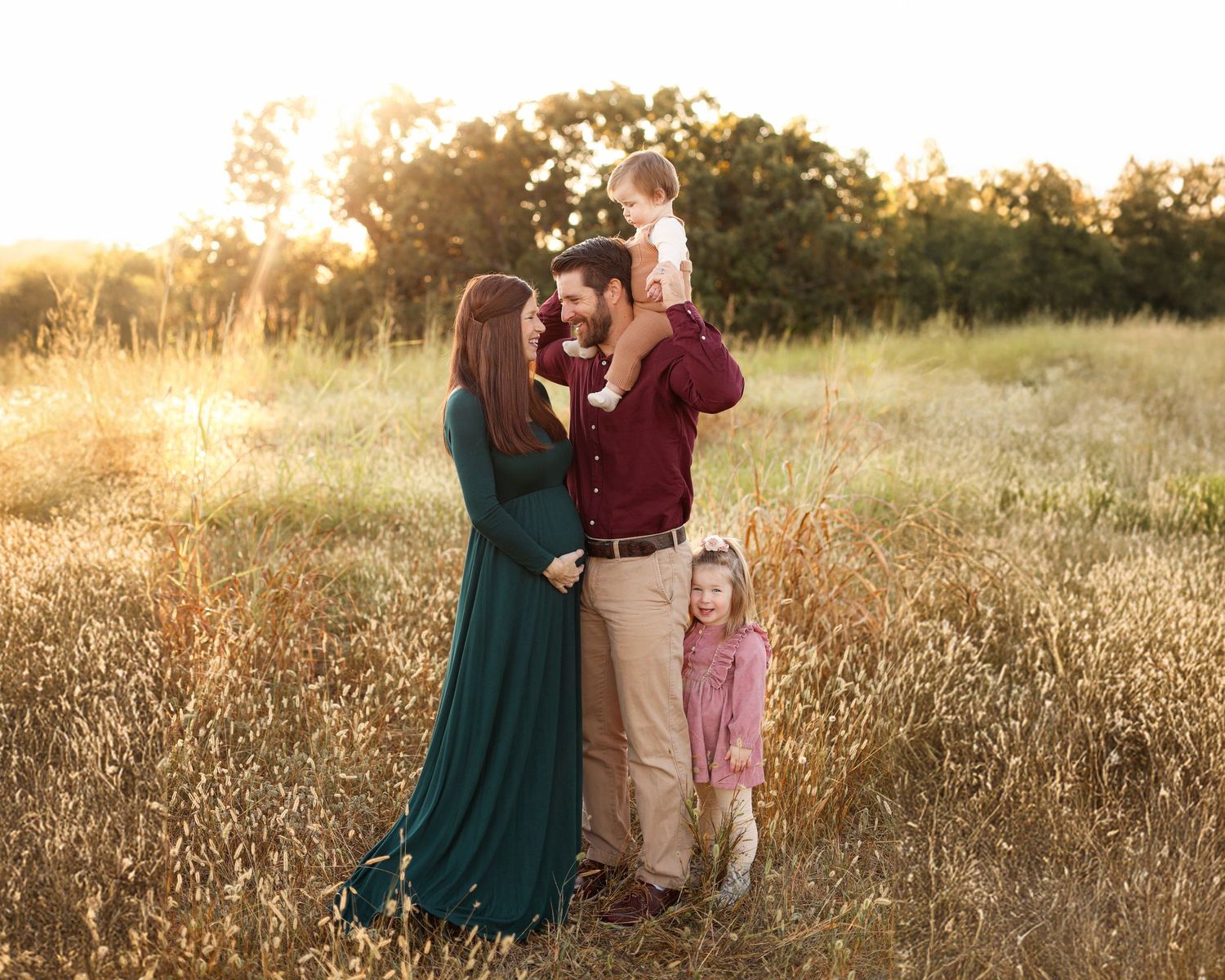 Family standing in a field with child on dads shoulders and a little girl smiling at the camera, mom is pregnant and smiling at dad