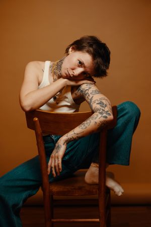 Artistic portrait of person with tattoos wearing white tank top and teal pants lounging on wooden chair against brown backdrop.