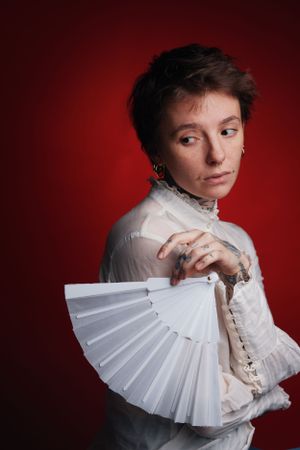 A dramatic portrait of a person in a vintage white high-collar blouse holding a decorative folding fan against a red background.
