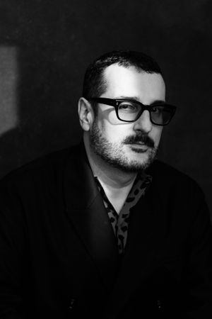 Black and white portrait of a bearded man wearing glasses against a dark background.
