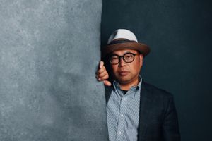 Contemporary businessman in a fedora and glasses leans against dark grey textured wall.