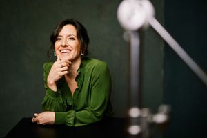 A woman in an emerald green blouse smiles warmly while resting her chin on her hand against a dark background.