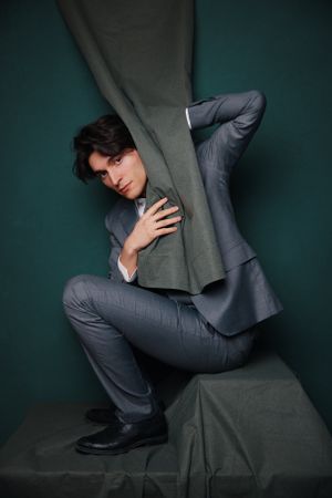 Fashion portrait of model interacting with grey fabric against dark green backdrop.