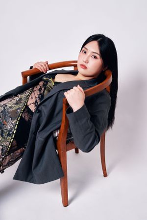 Fashion model poses artistically on wooden chair wearing black blazer with lace detail against white studio background.