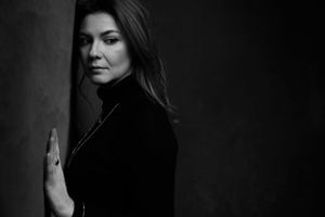 Dramatic black and white portrait against dark background shows person in black turtleneck with long dark hair.