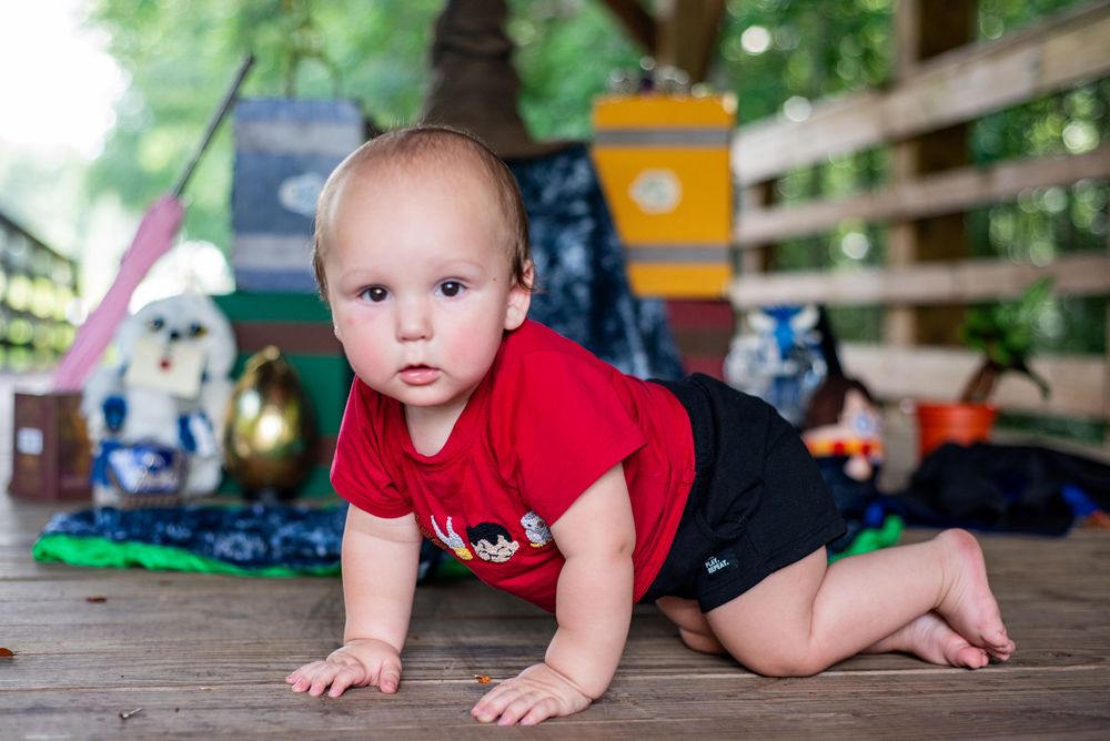 Baby in red shirt and dark shorts crawls on wooden deck with toys visible in background.