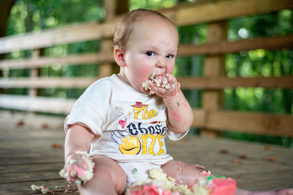 Baby in white onesie eating cake with fingers and making a mess during first birthday celebration outdoors.