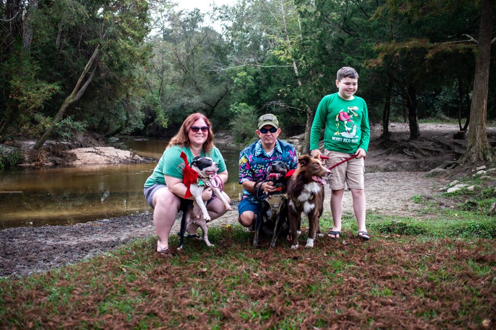 Group in matching outfits poses with their dogs by a creek in a wooded outdoor setting during summer.