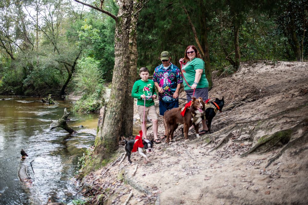 Group with dogs standing on a sloping dirt riverbank surrounded by trees and greenery.