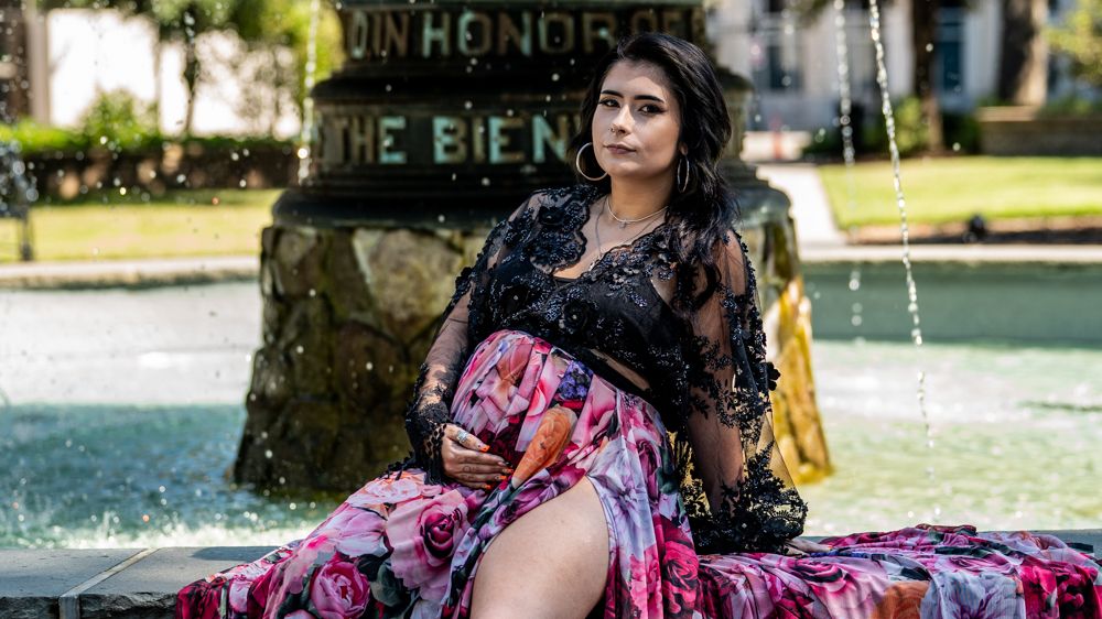 A person in a floral dress poses elegantly near a water fountain in a garden setting.