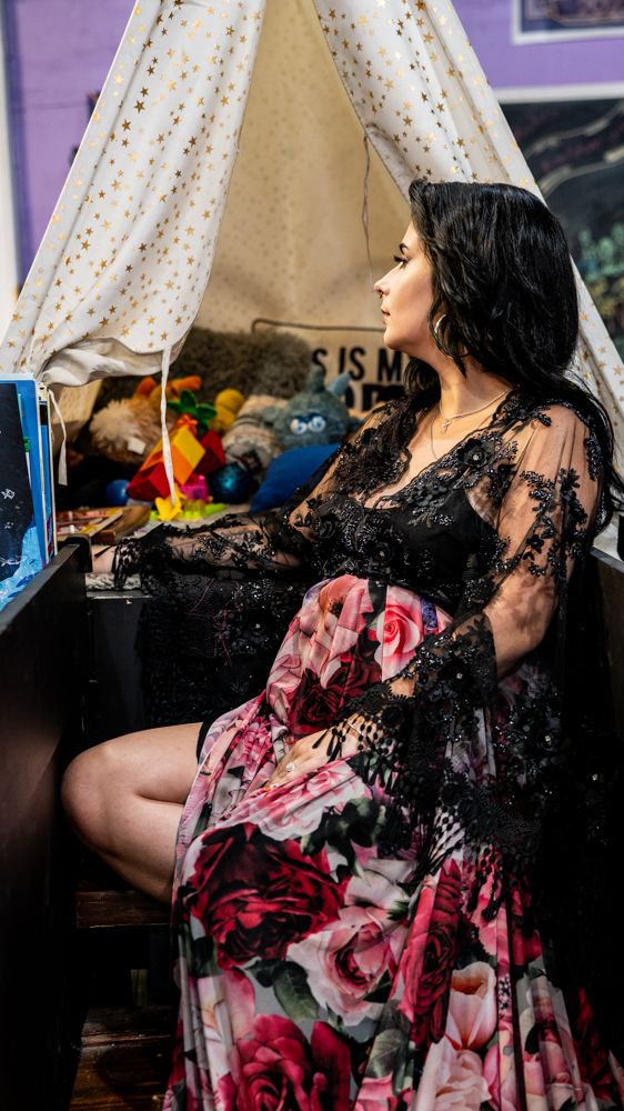 A person in a floral dress sits against a purple wall with white curtains in a casual indoor setting.