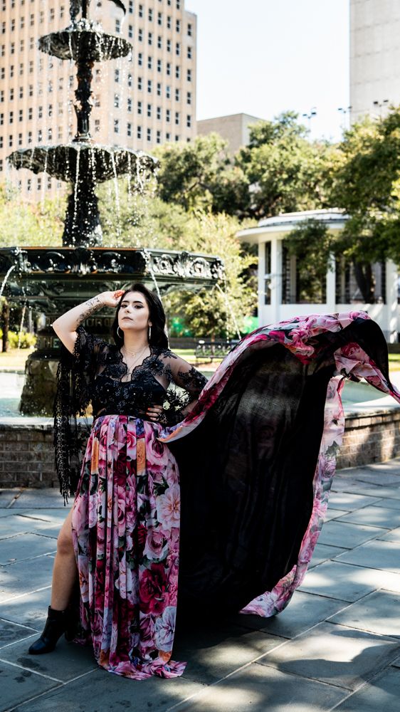 A figure in a flowing black and pink floral dress poses dramatically in front of an urban fountain on a sunny day.