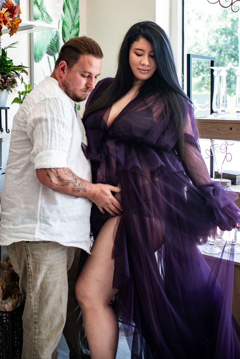A couple in white and purple outfits dance together in a bright sunlit room with large windows.