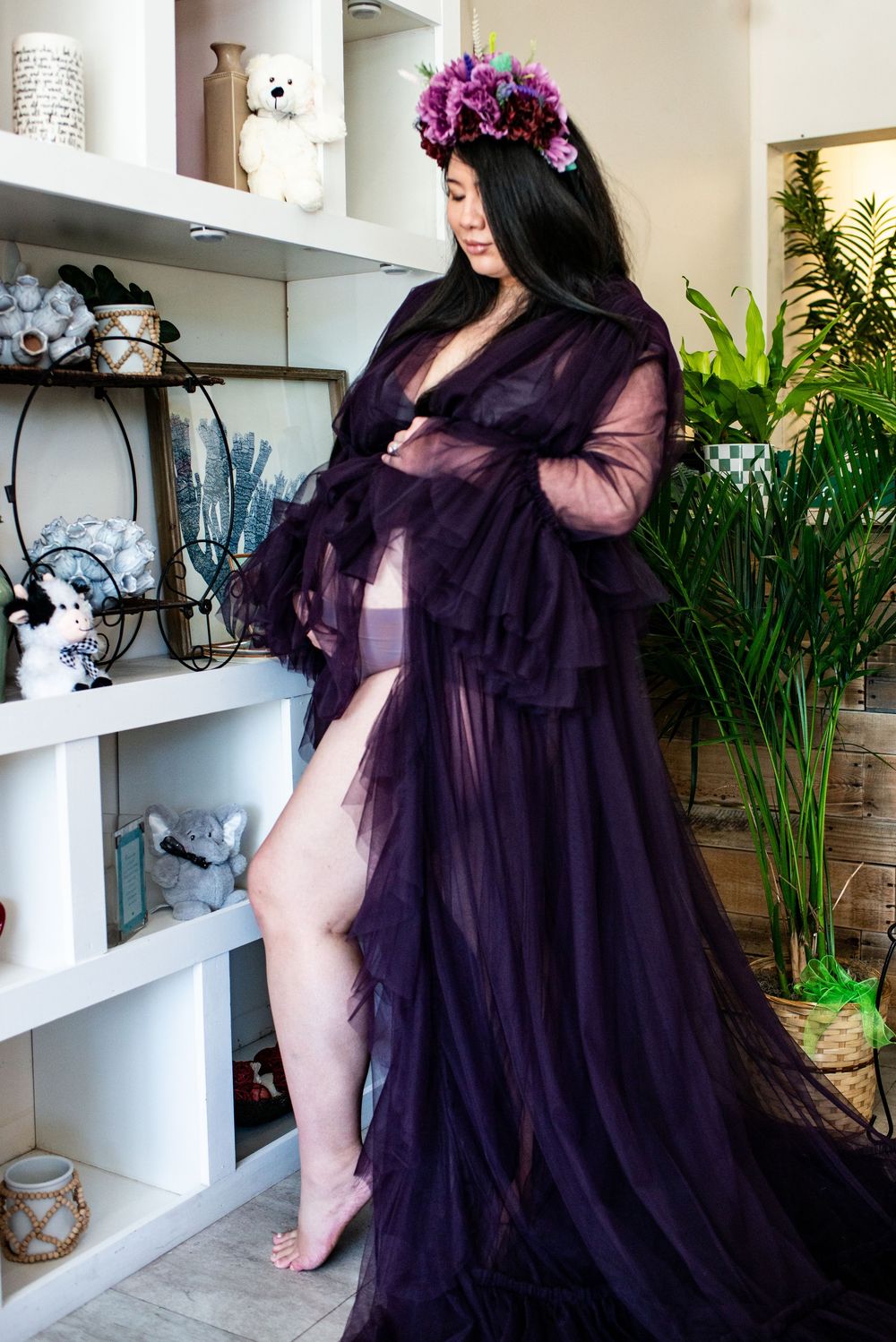 A flowing purple sheer maternity gown with ruffled sleeves is showcased in a bright indoor setting with plants.