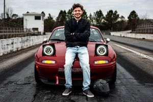 Someone sits on the hood of a red Mazda Miata sports car at a racetrack with pop-up headlights visible.