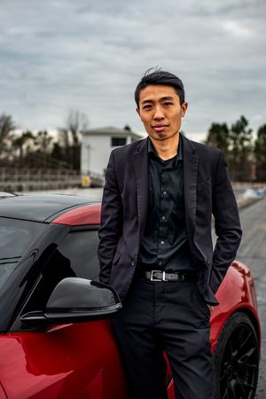 A person in a black suit poses next to a sleek red sports car on an overcast day.