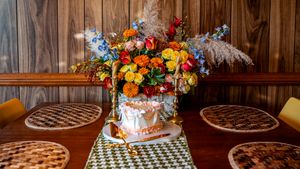 Festive birthday cake with colorful floral arrangement surrounded by pies on a wooden table with checkered runner.