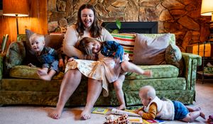 Woman sitting on green sofa laughing with two toddlers and a dog in a cozy living room.
