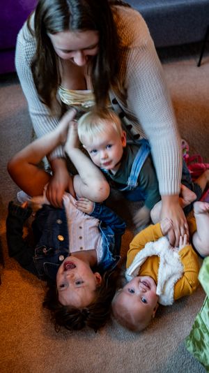 A young woman sits on the floor playing with three small children, laughing and cuddling together in a cozy living room.