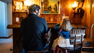 A man and young girl sit together at a wooden bar counter in a rustic wood-paneled room with artwork on the wall.