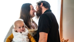 A couple shares a tender kiss while holding their adorable baby dressed in a white fluffy outfit indoors.