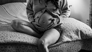 Black and white photo of a pregnant woman sitting on a bed, cradling her bare baby bump with both hands.