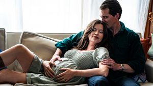 Pregnant woman resting on couch with partner, both smiling and cradling her baby bump in a cozy living room.
