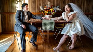 Bride and groom sit apart at a wooden table, looking distant, in a rustic room with floral centerpiece.