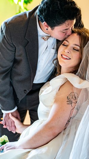 Bride smiling joyfully as groom kisses her forehead, bride wearing white gown and veil with floral arm tattoo.