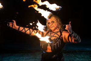 A fire performer in dark gothic attire dances with flaming torches against a dark background.