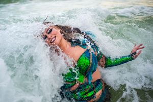 A performer in a green sequined costume dances in splashing ocean waves.