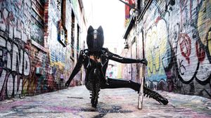 Person in black latex costume and cat ears poses dramatically in a graffiti-covered urban alley.