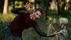A person in a burgundy sweater playfully interacts with a plastic skeleton decoration in an outdoor setting.