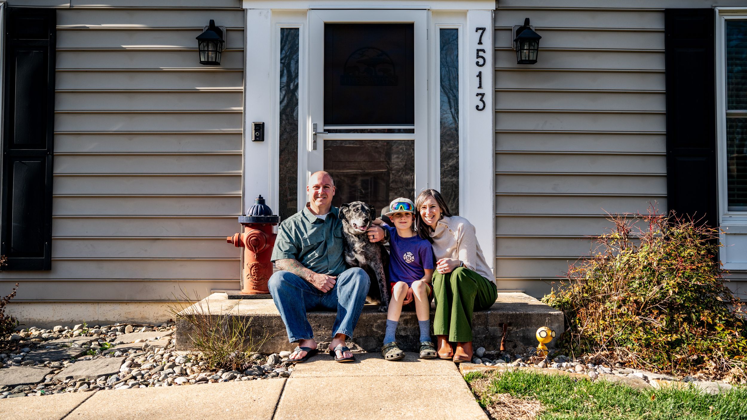 Family with dog posing happily on front porch steps of home numbered 7513 on a sunny day.