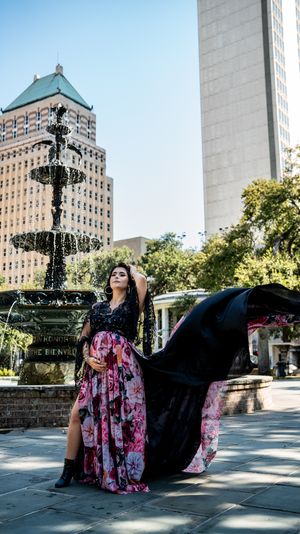 A person in a floral dress and black cape poses dramatically in front of an ornate fountain in an urban plaza.