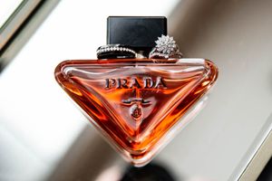A triangular Prada perfume bottle with amber-colored liquid displayed on a surface with wedding rings.