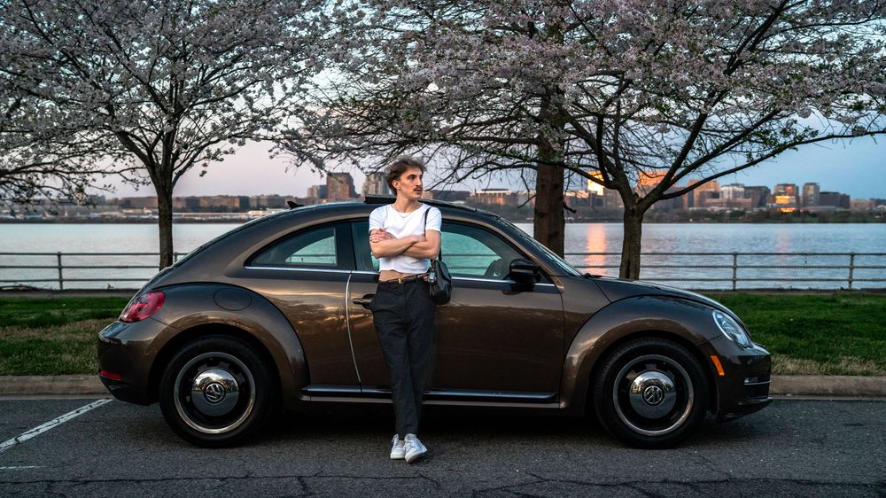 Person leaning against a brown Volkswagen Beetle near cherry blossom trees by a waterfront at dusk.