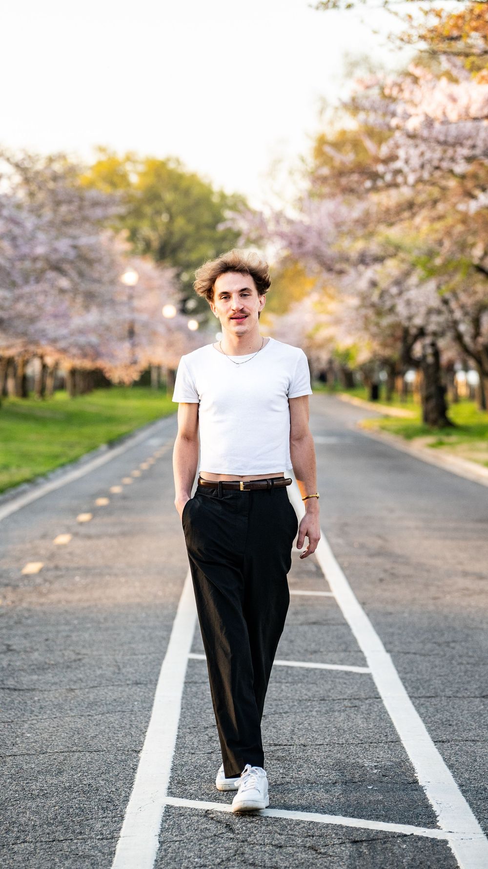 Young man in white crop top and black trousers walking on a road lined with cherry blossom trees in spring.