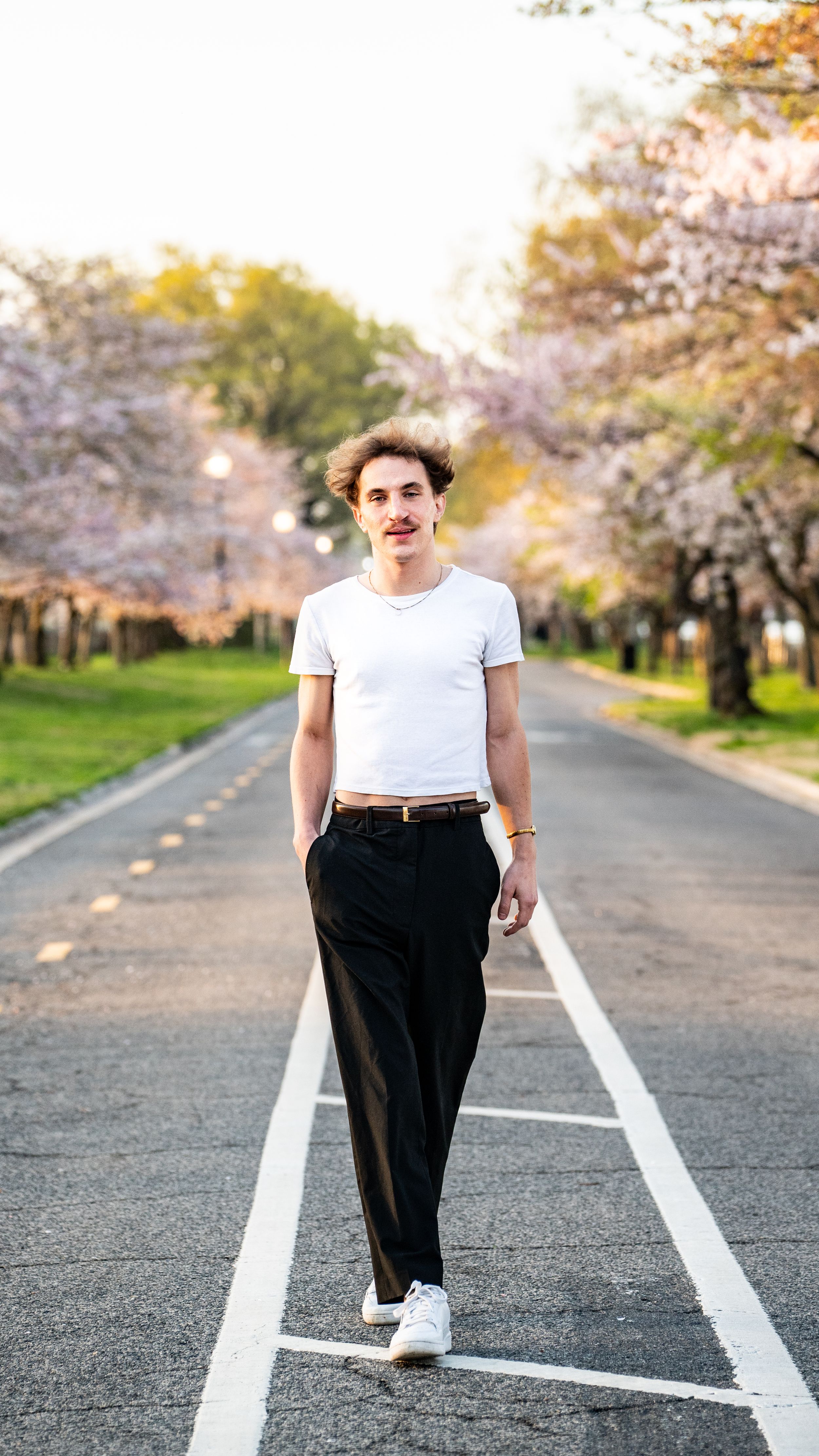 Young man in white crop top and black trousers walking on a road lined with cherry blossom trees in spring.