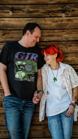 A couple in casual wear leans against a rustic wooden wall sharing candid laughing moments together.