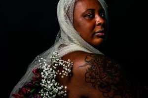 A dramatic portrait against a dark background features delicate baby's breath flowers and intricate shoulder tattoos.