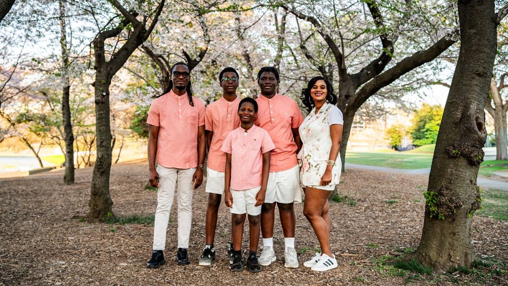 Family of five poses among cherry blossom trees wearing coordinated pink and white outfits in a park.