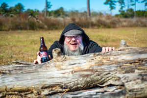 Someone in a black hat peers over a fallen log with a beer bottle in a sunny outdoor setting.