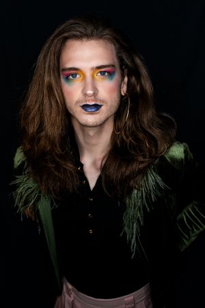 A dark artistic portrait featuring dramatic makeup with blue lipstick, colorful eyeshadow, and a black jacket accented with green feathers.