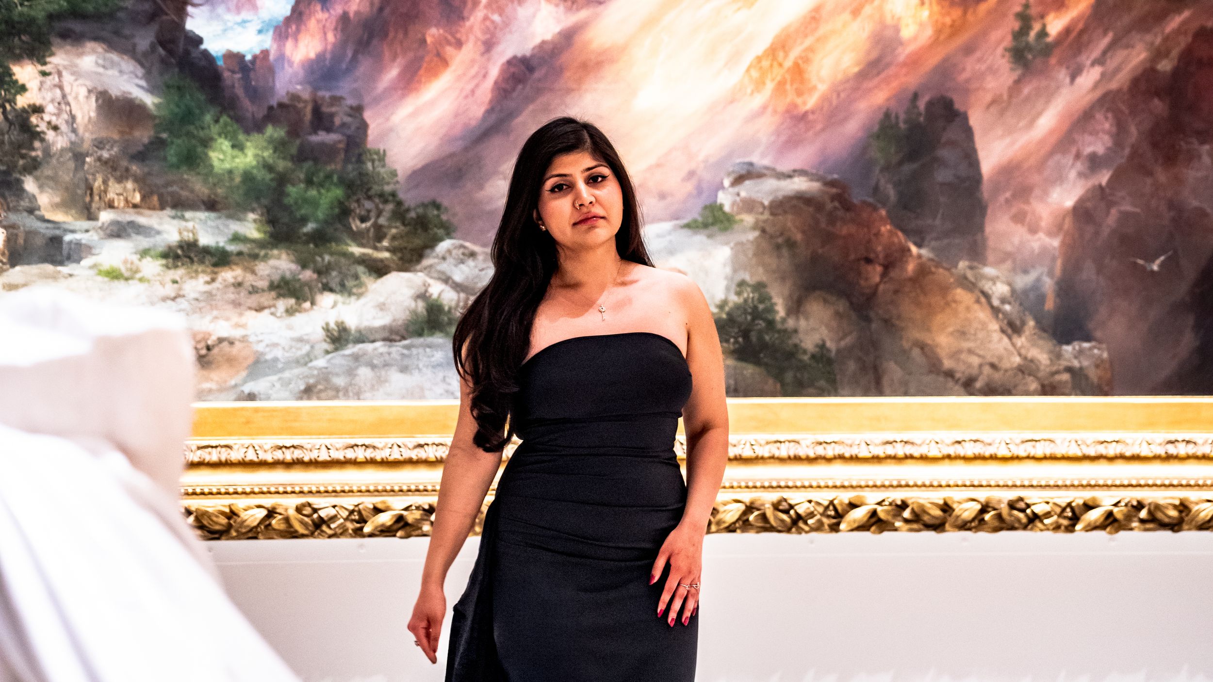 Woman in black strapless dress poses elegantly in front of a large colorful mountain landscape painting.