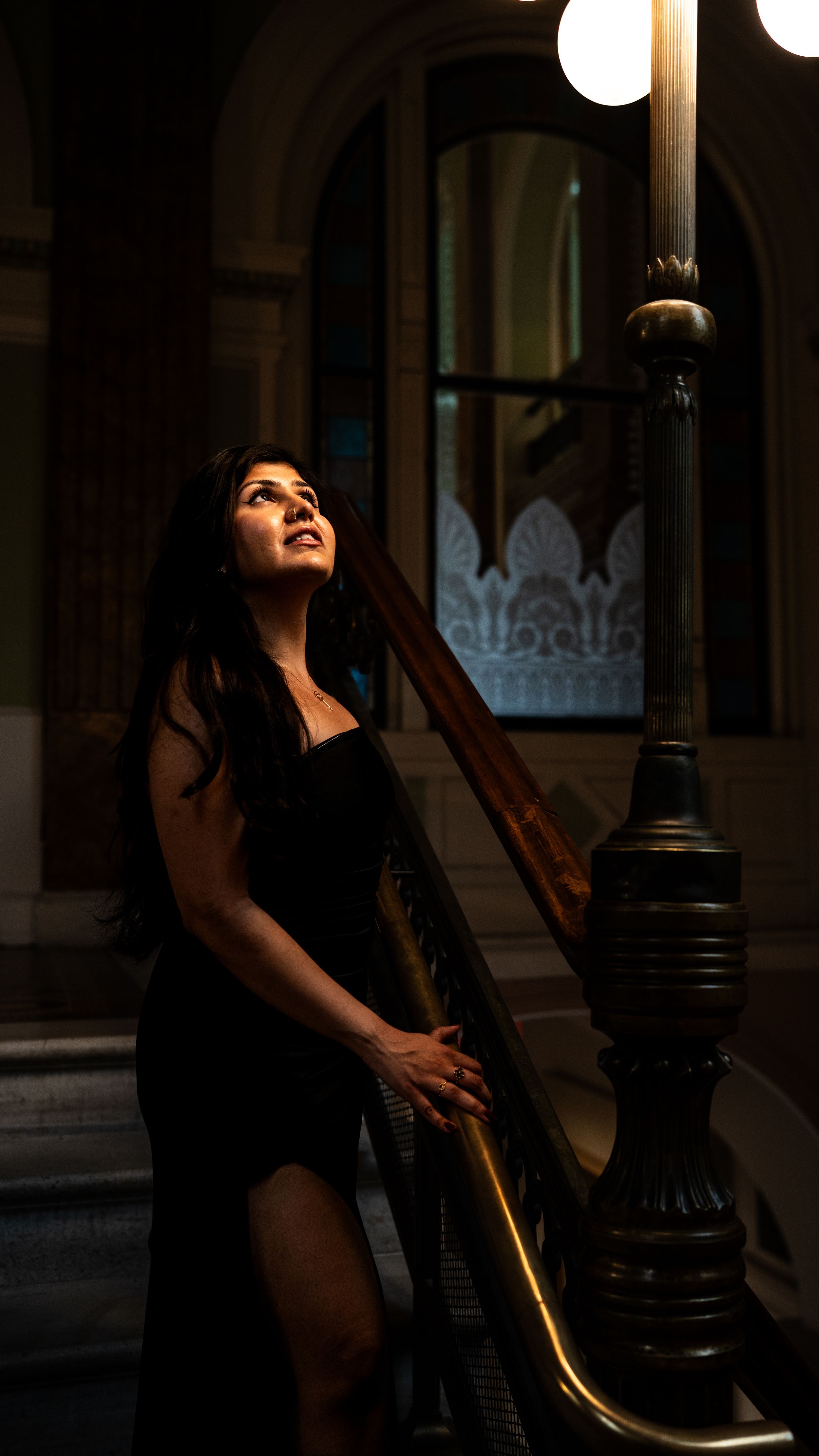 Woman in black dress gazes upward beside ornate lamp post on elegant staircase in dimly lit historic building.