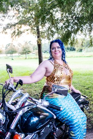 A person in a mermaid costume with blue hair sits on a motorcycle under a tree in a park setting.